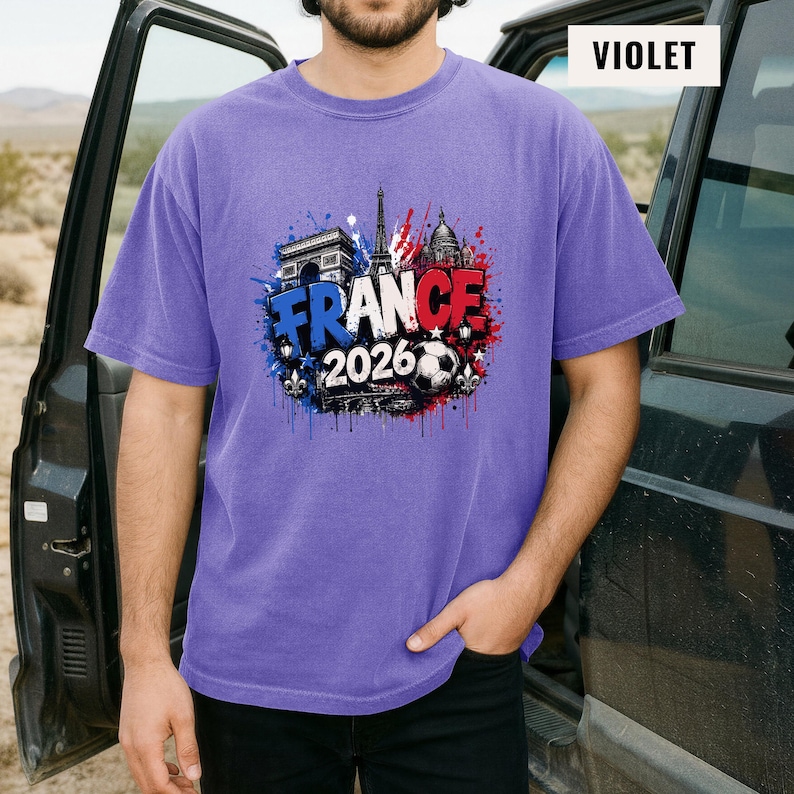 May include: Violet short-sleeve t-shirt featuring a graphic design with the text "FRANCE 2026" in white, blue, and red, along with images of the Eiffel Tower, Arc de Triomphe, and a soccer ball. The shirt is a solid violet color.