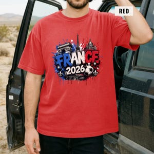 May include: Red t-shirt with a graphic design featuring the word "FRANCE" in blue and white, the year "2026", and images of the Eiffel Tower, Arc de Triomphe, and a soccer ball. The shirt is short-sleeved.