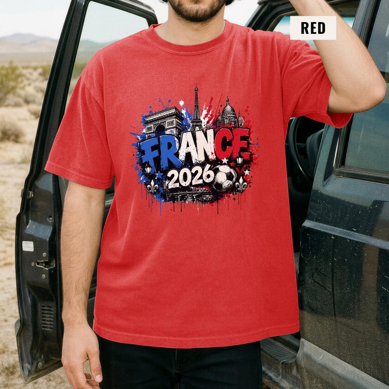 May include: Red t-shirt with a graphic design featuring the word "FRANCE" in blue and white, the year "2026", and images of the Eiffel Tower, Arc de Triomphe, and a soccer ball. The shirt is short-sleeved.