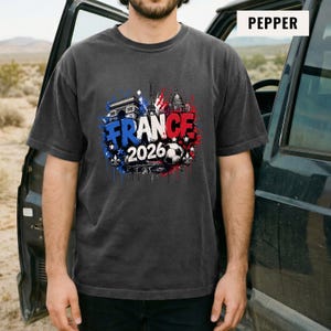 May include: Pepper-colored t-shirt featuring a graphic design with the word "FRANCE" in blue and red, the year "2026", and a soccer ball. The design includes illustrations of the Eiffel Tower, Arc de Triomphe, and other French landmarks.