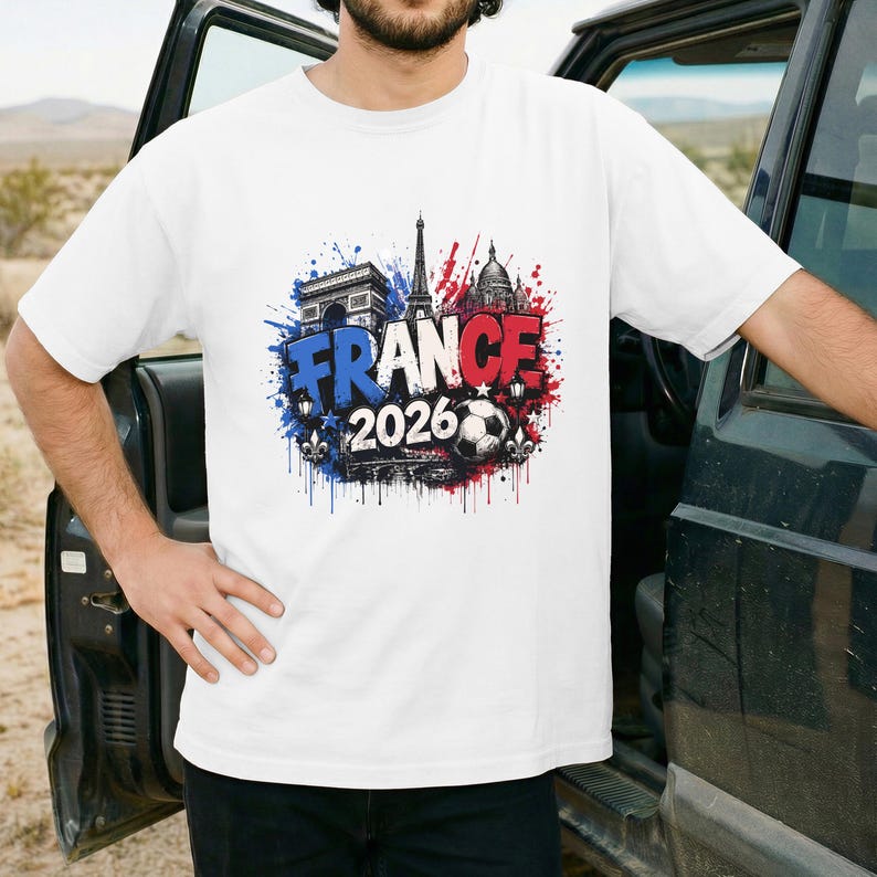 May include: White t-shirt featuring a colorful graphic design with the word "FRANCE" in blue and red, along with the year "2026" and a soccer ball. The design includes illustrations of the Eiffel Tower, Arc de Triomphe, and other landmarks.