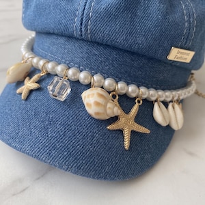 Op de afbeelding: Een blauwe denim pet versierd met parels en schelpen. De hoed heeft een decoratieve streng parels, schelpen, zeesterren en een heldere kristallen bedel. Een klein goudkleurig label leest "Ironmat Fashion".