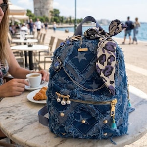 Peut inclure: Un sac à dos en jean bleu avec un motif patchwork, orné de clous dorés, d'une fermeture éclair et d'une écharpe décorative. Le sac présente un motif d'étoiles et est orné de breloques et d'un pompon. Le sac à dos est posé sur une table en marbre.