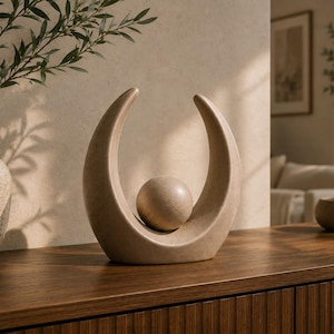 Pode incluir: Uma escultura de cerâmica bege com formato de meia-lua que envolve uma esfera central. O objeto possui um acabamento liso e fosco, com um design curvo e simétrico. É apresentado como uma peça decorativa independente com base sólida.