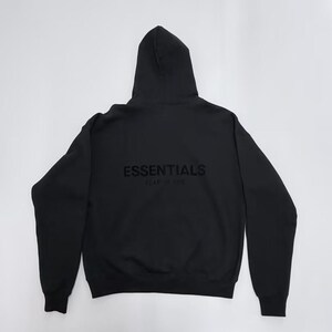 May include: Black hoodie with the word "ESSENTIALS" printed on the back. The hoodie has a hood and long sleeves. The text "FEAR OF GOD" is printed below "ESSENTIALS".