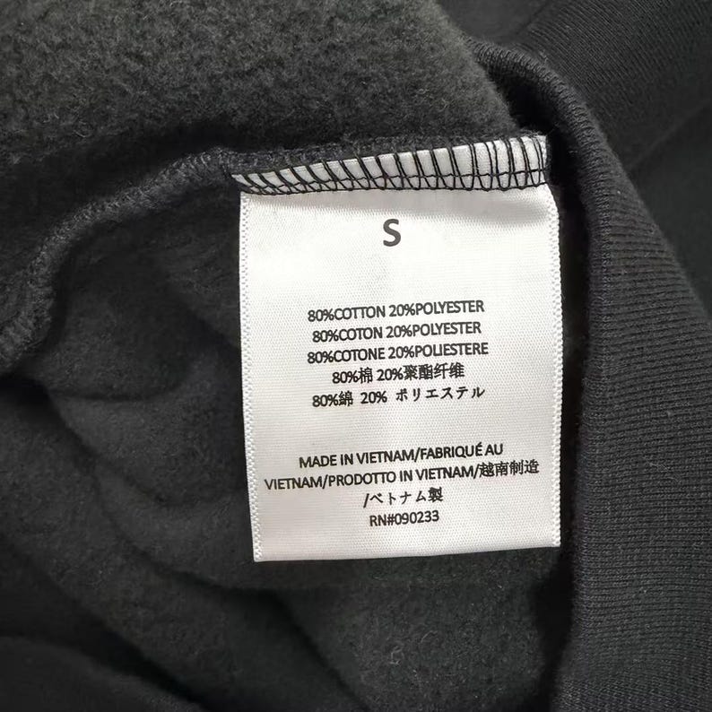 May include: Close-up of a black garment tag with the letter "S" at the top. The tag lists the fabric composition as 80% cotton and 20% polyester in multiple languages. The tag also indicates the item was made in Vietnam.