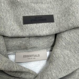 May include: A gray hoodie with the word "ESSENTIALS" on a rectangular patch at the hood and on a tag at the neckline. The hoodie is made of a soft, textured fabric.