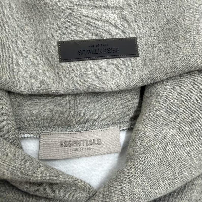 May include: A gray hoodie with the word "ESSENTIALS" on a rectangular patch at the hood and on a tag at the neckline. The hoodie is made of a soft, textured fabric.