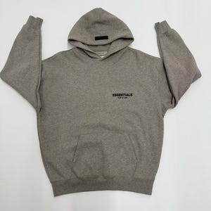May include: A gray hoodie with a hood and a front pocket. The word "ESSENTIALS" is printed in black on the front. The hoodie has long sleeves and a relaxed fit. The hoodie is made of a soft, comfortable material.