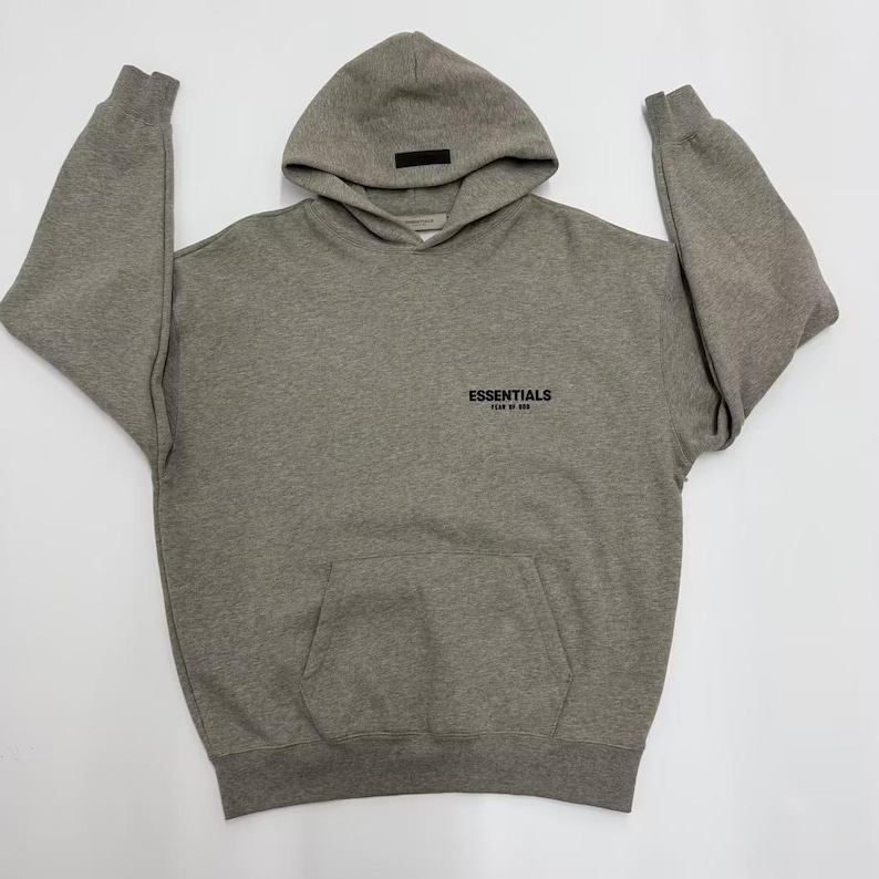 May include: A gray hoodie with a hood and a front pocket. The word "ESSENTIALS" is printed in black on the front. The hoodie has long sleeves and a relaxed fit. The hoodie is made of a soft, comfortable material.