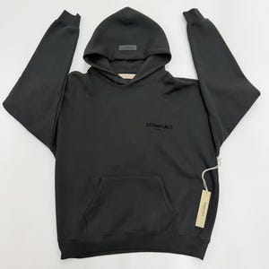 May include: Black hoodie with a hood and a front pocket. The word "ESSENTIALS" is printed on the front. A small tag is attached to the side of the hoodie.