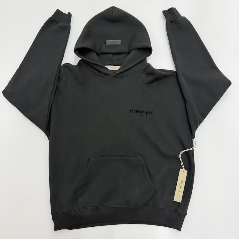 May include: Black hoodie with a hood and a front pocket. The word "ESSENTIALS" is printed on the front. A small tag is attached to the side of the hoodie.