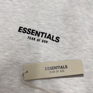 May include: Close-up of a light gray t-shirt with the word "ESSENTIALS" printed in black, above "FEAR OF GOD." A rectangular tag with the same text hangs below the shirt.