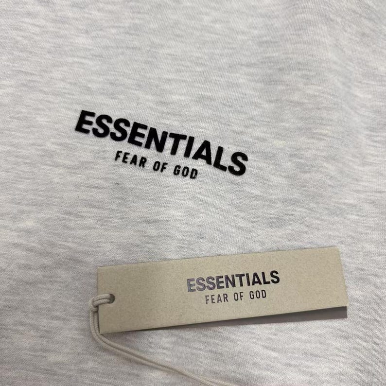 May include: Close-up of a light gray t-shirt with the word "ESSENTIALS" printed in black, above "FEAR OF GOD." A rectangular tag with the same text hangs below the shirt.