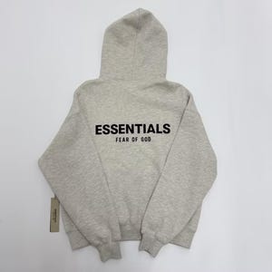 May include: A light gray hoodie with a hood and long sleeves. The word "ESSENTIALS" is printed in black block letters across the back, with "FEAR OF GOD" below it. A tag is attached to the bottom left.