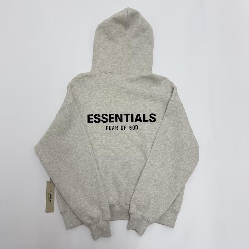 May include: A light gray hoodie with a hood and long sleeves. The word "ESSENTIALS" is printed in black block letters across the back, with "FEAR OF GOD" below it. A tag is attached to the bottom left.