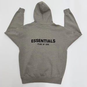 May include: A gray hoodie with a hood and long sleeves. The word "ESSENTIALS" is printed in black block letters across the back, with "FEAR OF GOD" printed below it. The hoodie is made of a soft, comfortable material.