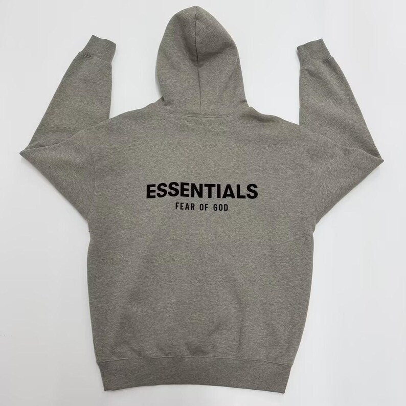 May include: A gray hoodie with a hood and long sleeves. The word "ESSENTIALS" is printed in black block letters across the back, with "FEAR OF GOD" printed below it. The hoodie is made of a soft, comfortable material.