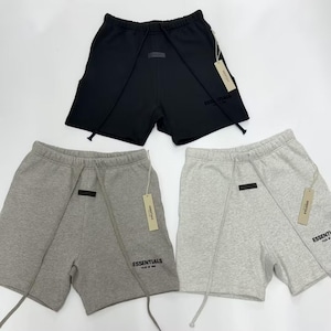 May include: Three pairs of athletic shorts in black, light gray, and medium gray. Each pair has a drawstring, a small rectangular patch, and a tag that reads "ESSENTIALS". The shorts are made of a soft material.