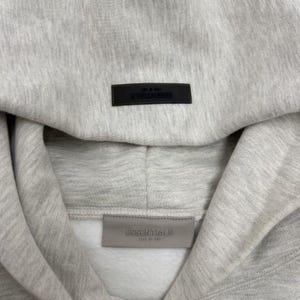 May include: Close-up of a light gray hoodie with a hood. The word "ESSENTIALS" is printed on a black rectangular patch at the top and a gray patch at the bottom. The hoodie is made of a soft, textured fabric.
