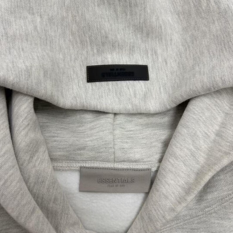 May include: Close-up of a light gray hoodie with a hood. The word "ESSENTIALS" is printed on a black rectangular patch at the top and a gray patch at the bottom. The hoodie is made of a soft, textured fabric.
