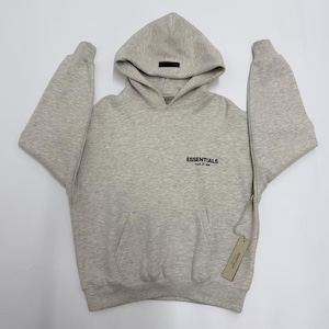 May include: A light gray hoodie with a drawstring hood and a kangaroo pocket. The word "ESSENTIALS" is printed in black on the chest. A black rectangular tag is on the hood, and a product tag hangs from the side.