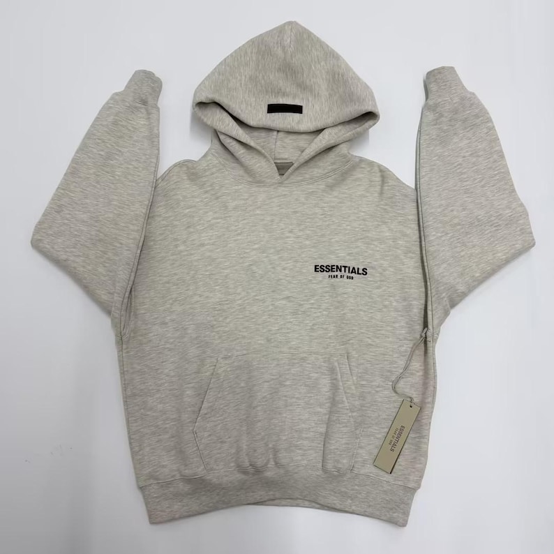 May include: A light gray hoodie with a drawstring hood and a kangaroo pocket. The word "ESSENTIALS" is printed in black on the chest. A black rectangular tag is on the hood, and a product tag hangs from the side.