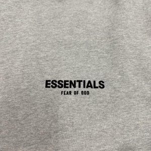 May include: Close-up of a heather gray sweatshirt with the word "ESSENTIALS" printed in black, with "FEAR OF GOD" printed below it. The fabric has a textured appearance.