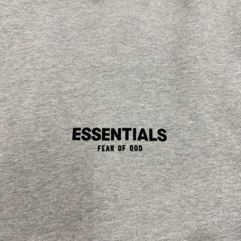 May include: Close-up of a heather gray sweatshirt with the word "ESSENTIALS" printed in black, with "FEAR OF GOD" printed below it. The fabric has a textured appearance.