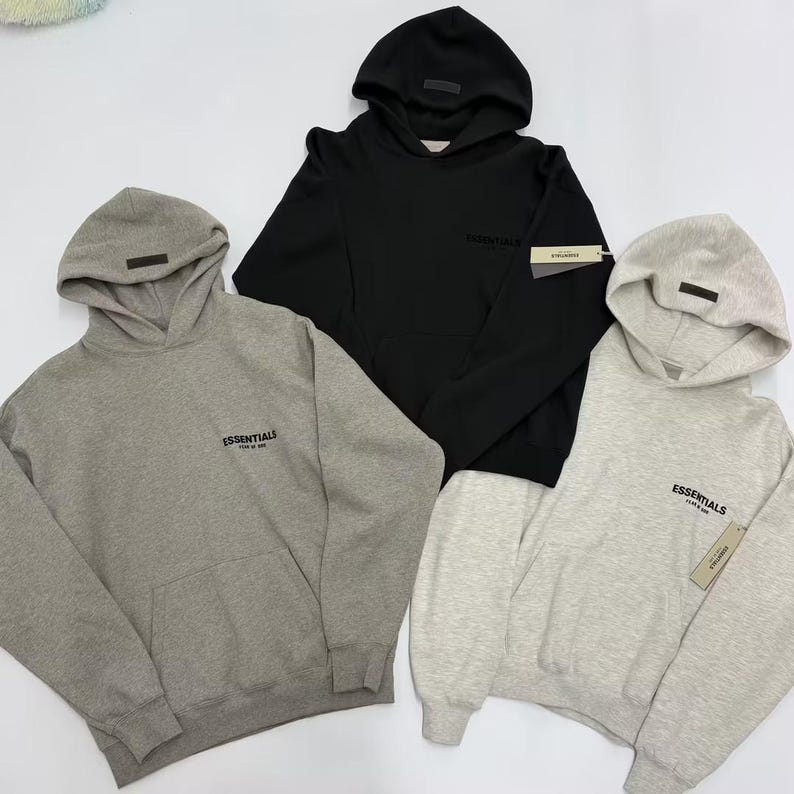 May include: Three hooded sweatshirts in gray, black, and light gray. Each sweatshirt has a hood, a small rectangular tag on the hood, and the word "Essentials" printed on the chest. One light gray sweatshirt has a tag attached to the sleeve.