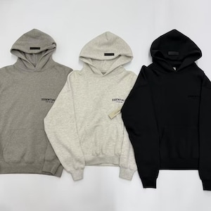 May include: Three hooded sweatshirts in gray, light gray, and black. Each sweatshirt has a front pocket, a hood with a small rectangular patch, and the word "ESSENTIAL" printed on the chest. The light gray sweatshirt has a tag.