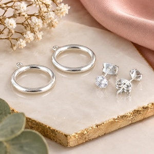 May include: Two silver-toned circular jewelry components with small loops at the top, accompanied by a pair of silver-toned stud earrings featuring clear, round-cut stones and matching metal butterfly backings.