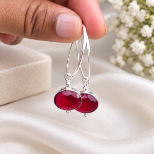 May include: A pair of silver hoop earrings featuring faceted, oval-shaped deep red gemstones suspended from the bottom of each hoop. The earrings have a polished metallic finish and a secure wire closure mechanism, showcasing a vibrant crimson hue.