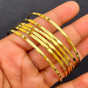 Op de afbeelding: Een set van vijf dunne, goudkleurige metalen armbanden met een gehamerd, getextureerd oppervlak. De armbanden zijn cirkelvormig en hebben een reflecterende, gepolijste afwerking die het licht vangt, wat zorgt voor subtiele inkepingen op de banden.