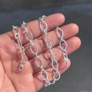 May include: A silver chain necklace featuring oval and circular links. The oval links have a textured design, and the necklace has a clasp. The jewellery is shown against a plain backdrop.