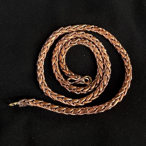 Puede incluir: Un collar de cadena de color cobre que presenta un diseño de eslabones trenzados y tejidos. El collar está enrollado en forma circular e incluye un pequeño mecanismo de cierre simple tipo gancho en el centro para asegurar los extremos de la cadena.