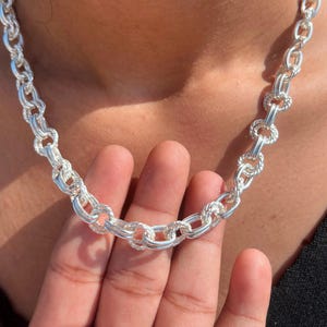 Peut inclure: Un collier à chaîne en argent composé d'une série de maillons circulaires entrelacés. Chaque maillon est formé d'un anneau extérieur lisse et poli associé à un anneau intérieur torsadé et texturé, créant un motif répétitif de cercles argentés.