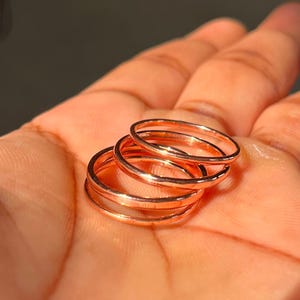 May include: A set of three thin, polished rose gold-toned metal rings. Each ring features a smooth, circular band with a reflective surface. The rings are stacked slightly overlapping each other, showcasing a consistent metallic copper-colored finish.