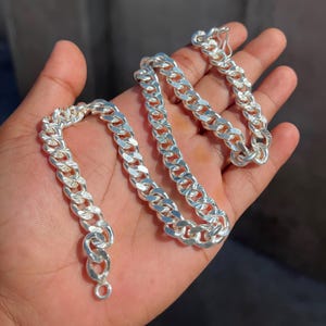 Puede incluir: Un collar de cadena de plata con un acabado pulido. La cadena se muestra en una mano, mostrando los eslabones y el cierre. Una pieza de joyería clásica.