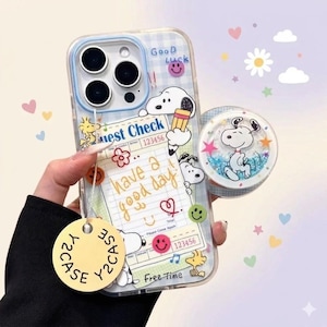 Puede incluir: Funda transparente para teléfono con un patrón de cuadros azules y gráficos de dibujos animados de Snoopy. La funda presenta un soporte emergente con Snoopy dentro de un círculo lleno de purpurina. El texto en la funda incluye "Good Luck" y "Have a good day!"