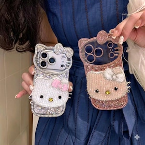 May include: Two decorative phone cases with a cartoon cat design. One is silver with a white cat face and a pink bow. The other is rose gold with a pink cat face and a white bow. Both cases are embellished with rhinestones.