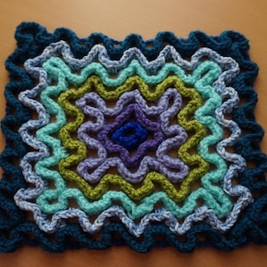 Könnte beinhalten: Ein quadratischer gehäkelter Untersetzer mit einem mehrfarbigen Design. Der Untersetzer ist aus blauem, grünem, violettem und weißem Garn gefertigt. Die Mitte des Untersetzers hat die Form eines Sterns.