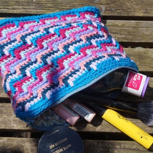 Op de afbeelding: Een blauwe gebreide tas met rits met een roze, blauwe en zwarte chevron-print. De tas is open en bevat make-upkwasten, een compact en een fles lotion met het label "No Rub Formula".