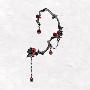 May include: A black ear cuff with a rose and thorn design. The cuff features red rose accents and dangling red teardrop-shaped gems. A chain with a teardrop gem hangs from the bottom of the cuff.