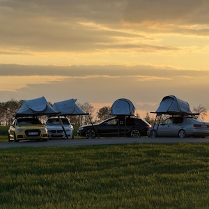 Könnte beinhalten: Vier Autos mit Dachzelten parken bei Sonnenuntergang auf einer Wiese. Die Autos sind in verschiedenen Farben gehalten, darunter Gelb, Silber und Schwarz. Die Zelte sind hellblau und scheinen zum Camping aufgebaut zu sein. Der Himmel ist eine Mischung aus Orange und Gelb.