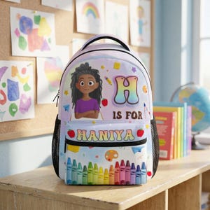 May include: A light-colored backpack featuring a cartoon illustration of a person with dark hair, the letter H, the text H IS FOR HANIYA, and a row of colorful crayons. The design includes small icons like a pencil, apple, and paint palette.
