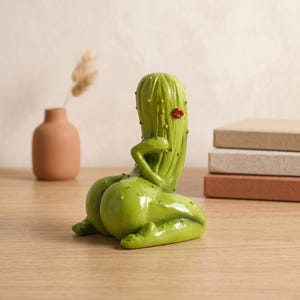 May include: A green cactus figurine shaped like a person in a kneeling pose. The cactus has a smooth, glossy finish with small raised bumps, a single arm resting on its hip, and bright red lips painted on the upper portion of the main body.