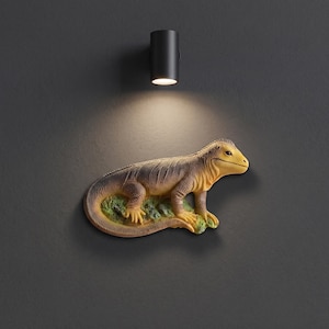 May include: A wall-mounted lizard sculpture featuring a textured body with shades of brown, tan, and yellow. The lizard is depicted in a standing position on a small, green, rocky base, with a black cylindrical light fixture positioned above it.
