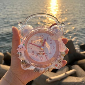 May include: A pink alarm clock with a clear handle and a mermaid tail design. The clock face has pink hands and numbers, with a floral design at the bottom. The clock is decorated with seashells and a small octopus figure.