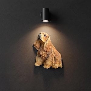 May include: A wall-mounted sculpture of a golden-brown spaniel dog with long, textured fur and floppy ears. The dog is depicted in a seated position, looking upward, and is illuminated by a small, cylindrical black light fixture mounted above.
